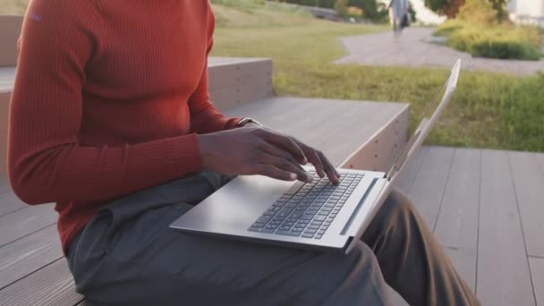 Midsectie slow mo shot van handen van onherkenbare Afro-Amerikaanse zakenman in slimme casualwear typen op laptop toetsenbord, zittend op trappen in het park in de zomer - Video