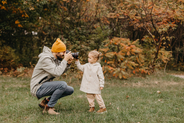 美しい秋の公園を歩きながら遊んでいる幸せな父親と娘。娘と週末の理想的な父親。父は娘の写真をプロのカメラで撮って - 写真・画像