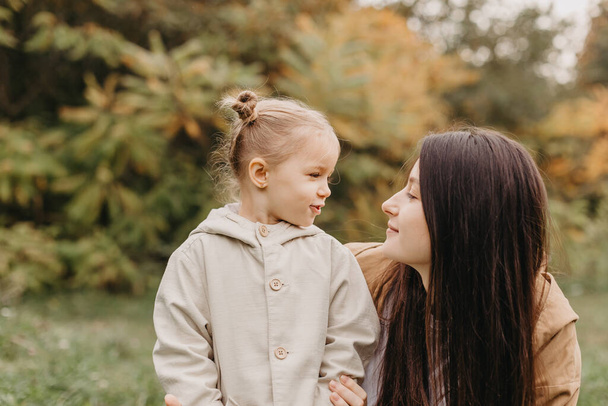 Young happy and smiling mom with her little daughter in arms hugging and kissing spending a weekend on a walk in autumn park. selective focus, noise effect, Autumnal mood - Photo, Image