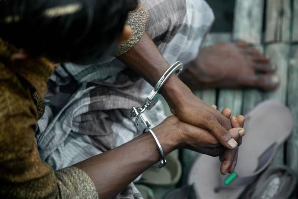 逮捕されたアジア系の犯罪者が手錠をかけられて座っている。若い村の被告人が上から座っているのが見えます。被告人の手に手錠をかける。犯罪者を逮捕. - 写真・画像
