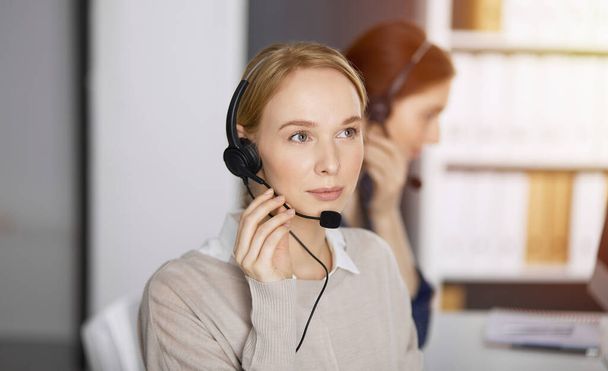 Friendly caucasian businesswoman talking by headset in sunny office. Call center and diverse people group in business - Valokuva, kuva