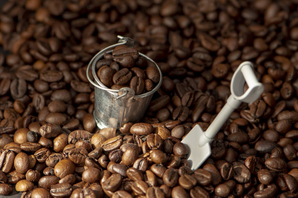 Een metalen emmer gevuld met koffiebonen en een kleine spatel tegen de achtergrond van verspreide koffie, selectieve focus. Concept: koffiemijn, koffiebonenoogst. - Foto, afbeelding