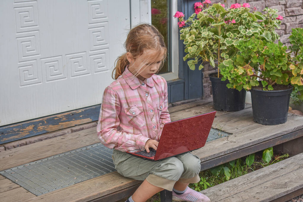 a girl child with a laptop is sitting on the porch of the house during a lesson on a remote photo without a filter - Photo, Image