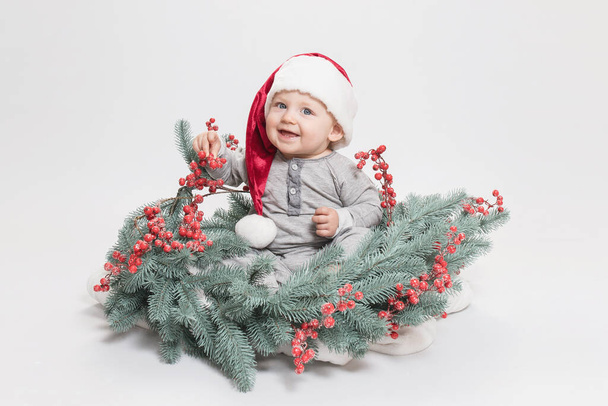 Adorabile bambino felice in un cappello di Babbo Natale seduto in ghirlanda decorativa da un albero di Natale. Faccia sorridente. Buon Natale, simpatico bambino allegro nel concetto di costume di Capodanno. Ricevuto. Bianco studio sfondo - Foto, immagini