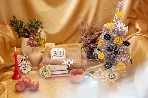 Uroczysta atmosfera imprezy z nowoczesnym drzewem Halloween. Przytulne dekoracje Halloween ze świecami, kubek pomarańczowy, suche kwiaty, domowe ciasteczka w kształcie dyni i ducha z książkami i kalendarzem.  - Zdjęcie, obraz