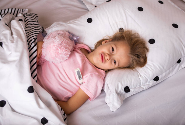 uma criança doente está na cama com um brinquedo macio e mede a temperatura com um termômetro - Foto, Imagem