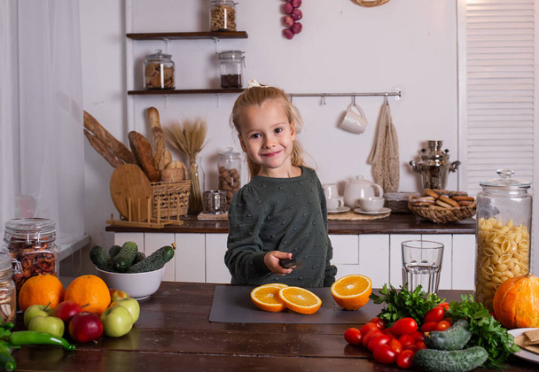 een blond meisje in een groene trui zit aan een tafel en snijdt een sinaasappel - Foto, afbeelding
