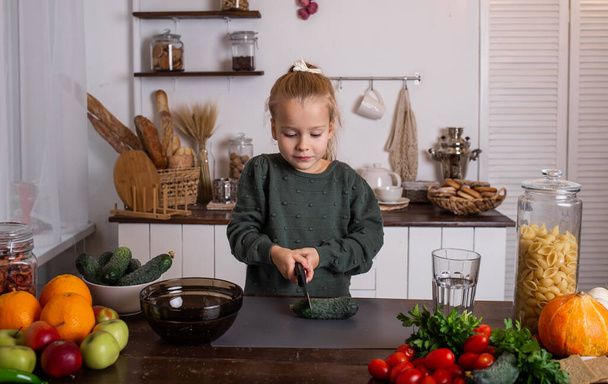Egy szőke kislány zöld pulcsiban ül az asztalnál, és uborkát vág. - Fotó, kép