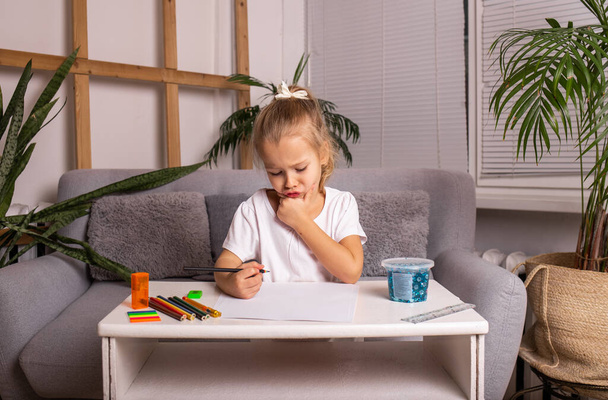 malá holčička v bílém tričku sedí u stolu a kreslí tužkou - Fotografie, Obrázek