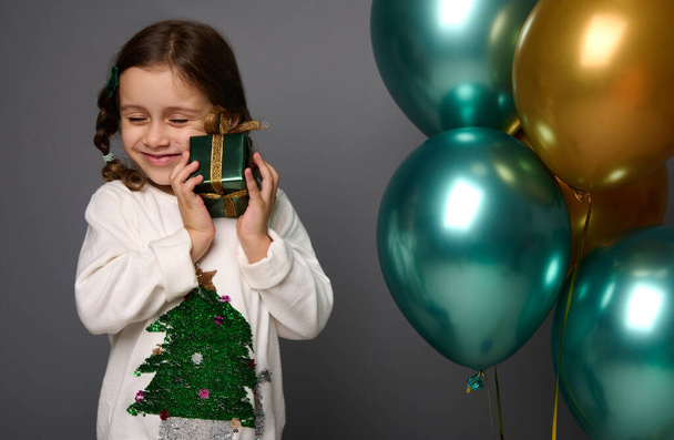 Adorable Caucasian little girl with closed eyes gently hugs her Christmas gift, standing near beautiful shiny air balloons of green metallic and golden colors, isolated portrait on gray background - 写真・画像