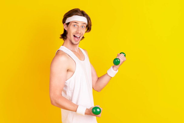 Profile side view portrait of attractive cheerful guy lifting small light dumbbell isolated over bright yellow color background - 写真・画像