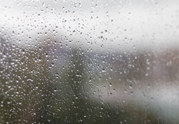 Jour de pluie à travers la fenêtre sur ciel gris nuageux et le fond des bâtiments de la ville. Concept. Paysage urbain du soir derrière la fenêtre en verre avec des gouttes d'eau qui coulent. - Photo, image