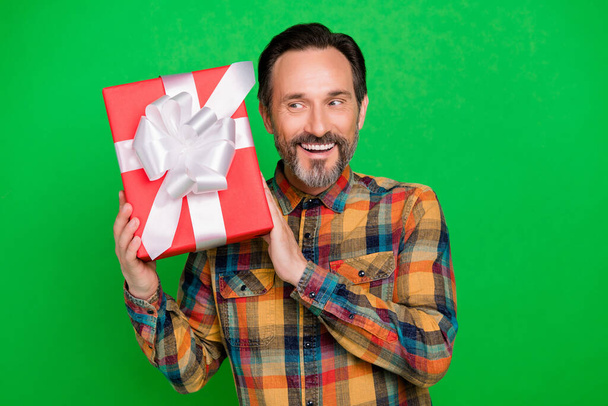 Portrait of attractive cheerful man curious holding in hands giftbox guessing what inside isolated over bright green color background - 写真・画像