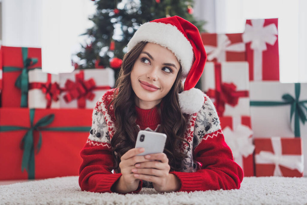 Photo of blogger lady lay carpet texting phone look up wear santa hat sweater in decorated home indoors - Zdjęcie, obraz