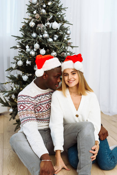 Portrait of smiling young couple sit near Christmas tree celebrate New Year together. Happy man and woman enjoy winter holiday celebration near decorated fir-tree - Foto, afbeelding
