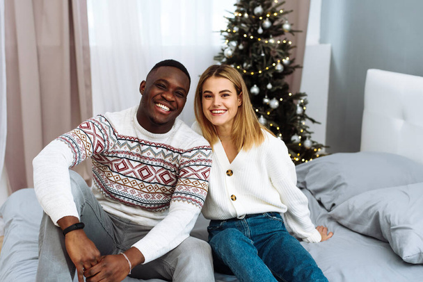 Birbirine aşık bir çift, bir Noel ağacının arka planında yan yana oturur ve kameraya gülümser. Noel ve Yeni Yıl kavramı - Fotoğraf, Görsel