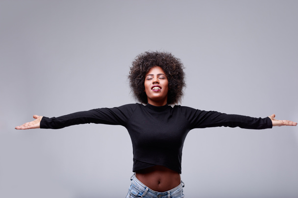 Genç siyahi kadın, kollarını açıp meditasyon yapmak için sessiz bir anın tadını çıkarıyor. Gri stüdyo arka planında, telif boşluğu olan sakin bir ifade var. - Fotoğraf, Görsel