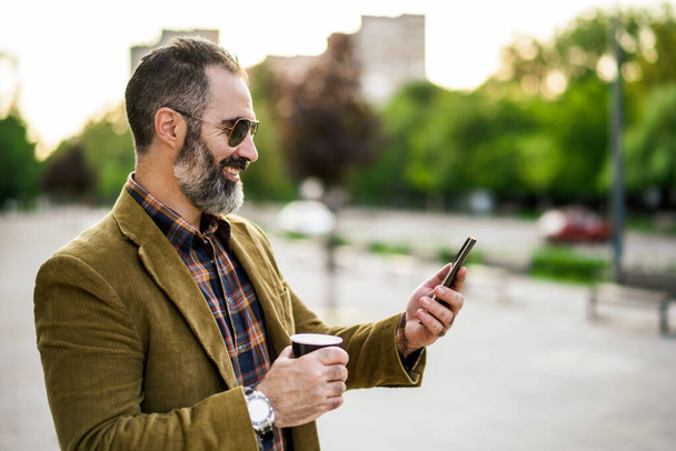 Portrait of modern businessman  with beard using mobile phone and drinking coffee while standing on the city street. - 写真・画像