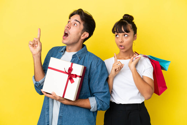 Young couple holding shopping bags and present isolated on yellow background thinking an idea pointing the finger up - Φωτογραφία, εικόνα