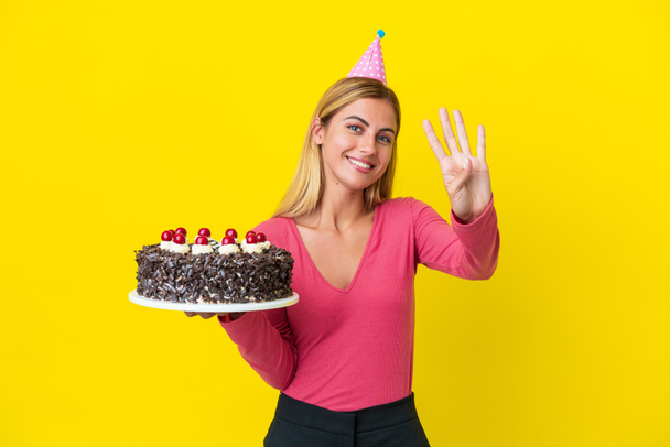 Blonde uruguayische Mädchen halten Geburtstagstorte isoliert auf gelbem Hintergrund glücklich und zählen vier mit den Fingern - Foto, Bild