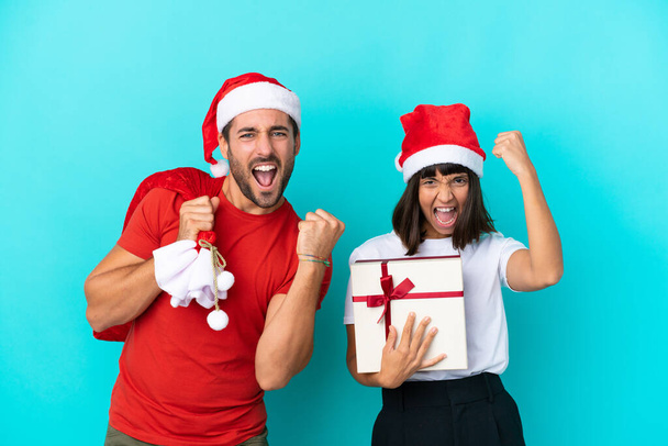 Giovane coppia con cappello di Natale distribuendo regali isolati su sfondo blu che celebrano una vittoria - Foto, immagini
