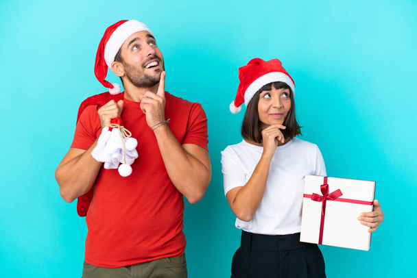 Νεαρό ζευγάρι με χριστουγεννιάτικο καπέλο που μοιράζει δώρα απομονωμένα σε μπλε φόντο σκεπτόμενο μια ιδέα ενώ κοιτάζει ψηλά - Φωτογραφία, εικόνα