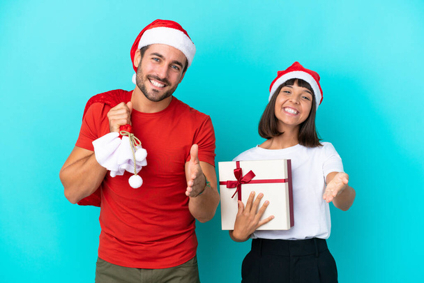 Junges Paar mit Weihnachtsmütze verteilt Geschenke auf blauem Hintergrund und schüttelt Hände, um ein gutes Geschäft abzuschließen - Foto, Bild