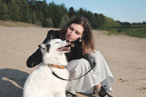 happy laika dog smiling and sitting next to its owner. young beautiful woman stroking her dog sitting on a sandy beach. togetherness and loving pets concept. - Photo, Image
