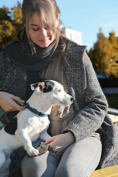 молодая женщина в пальто, обнимающая собаку, сидя на скамейке в осеннем парке. Джек Руссел терьер на коленях у женщины, смотрит в сторону. уход за животными, прогулки вместе и отдых на открытом воздухе. - Фото, изображение