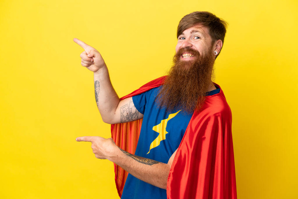 Redhead Super Hero άνθρωπος απομονώνονται σε κίτρινο φόντο δείχνοντας το δάχτυλο στο πλάι και παρουσιάζοντας ένα προϊόν - Φωτογραφία, εικόνα