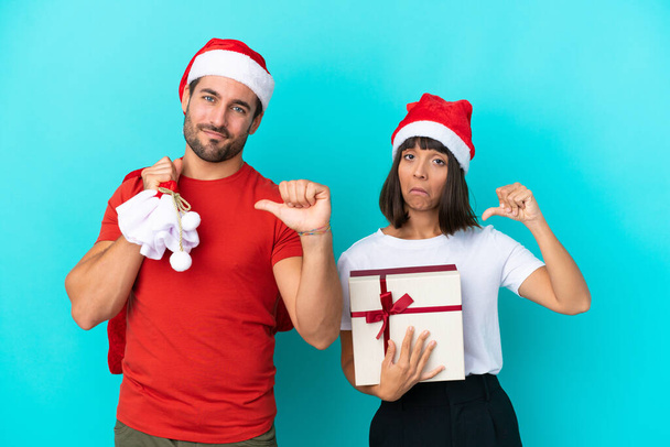 Mladý pár s vánočním kloboukem rozdávat dárky izolované na modrém pozadí hrdý a self-spokojen v lásce sami koncept - Fotografie, Obrázek