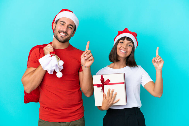 Noel şapkalı genç çift mavi arka planda izole edilmiş hediyeler dağıtıyor ve en iyinin işareti olarak parmağını kaldırıyor. - Fotoğraf, Görsel