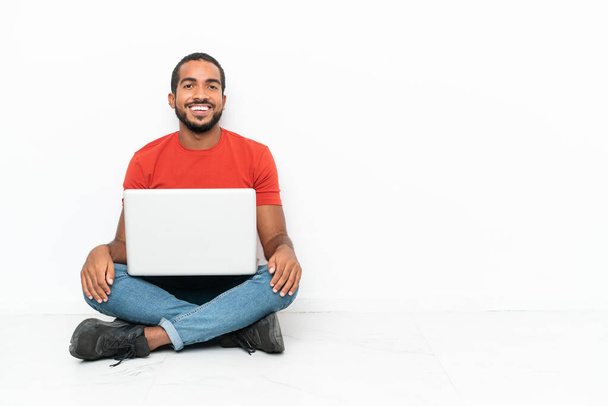 Junger ecuadorianischer Mann mit Laptop sitzt isoliert auf dem Boden vor weißem Hintergrund und lacht - Foto, Bild