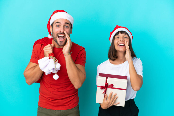 Fiatal pár karácsonyi kalapban ajándékokat osztogat elszigetelt kék háttérrel kiabálva és bejelentve valamit - Fotó, kép