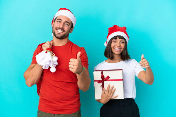 Молода пара з різдвяним капелюхом роздає подарунки ізольовані на синьому фоні, даючи великі пальці вгору жестом, тому що сталося щось хороше
 - Фото, зображення