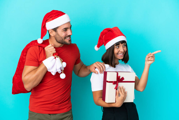 Giovane coppia con cappello di Natale distribuendo regali isolati su sfondo blu che presenta un'idea mentre guarda sorridente verso - Foto, immagini