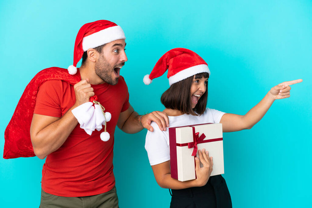 Νεαρό ζευγάρι με χριστουγεννιάτικο καπέλο μοιράζει δώρα που απομονώνονται σε μπλε φόντο δείχνοντας προς την πλευρά για να παρουσιάσει ένα προϊόν - Φωτογραφία, εικόνα