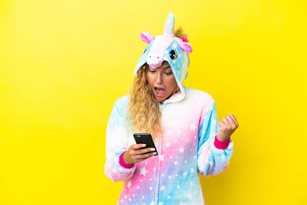 Chica con el pelo rizado con un pijama unicornio aislado sobre fondo amarillo sorprendió y envió un mensaje - Foto, Imagen