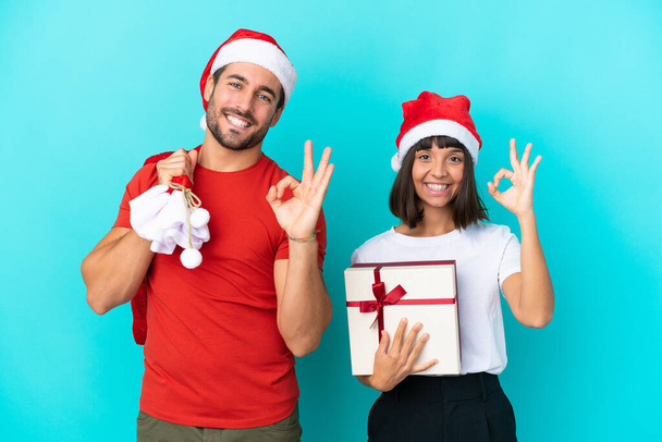 Νεαρό ζευγάρι με χριστουγεννιάτικο καπέλο μοιράζοντας δώρα που απομονώνονται σε μπλε φόντο δείχνει μια πινακίδα εντάξει με τα δάχτυλα - Φωτογραφία, εικόνα