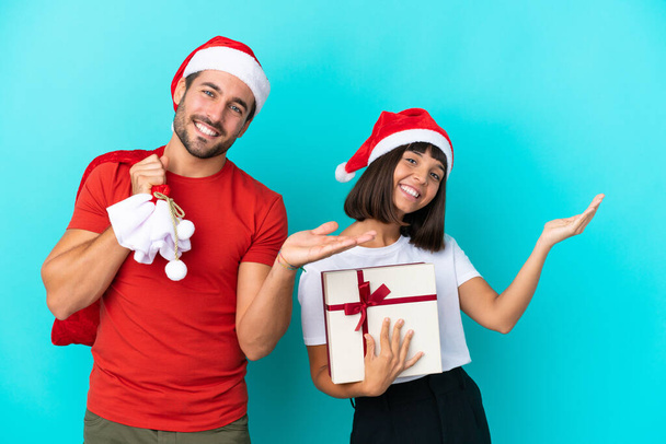 Νεαρό ζευγάρι με χριστουγεννιάτικο καπέλο μοιράζει δώρα απομονωμένα σε μπλε φόντο απλώνοντας τα χέρια στο πλάι για την πρόσκληση να έρθει - Φωτογραφία, εικόνα