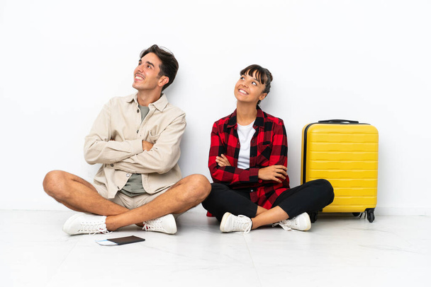 Young mixed race travelers couple sitting on the floor isolated on white background looking up while smiling - Foto, Imagen