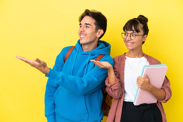 Mladý studentský pár izolovaný na žlutém pozadí ukazuje zpět a prezentuje produkt - Fotografie, Obrázek