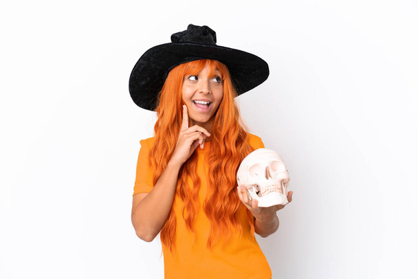 Молодая женщина смешанной расы, замаскированная под ведьму, держащая череп изолирован на белом фоне, думая об идее, глядя вверх - Фото, изображение