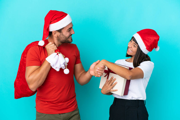 Νεαρό ζευγάρι με χριστουγεννιάτικο καπέλο μοιράζοντας δώρα που απομονώνονται στο μπλε φόντο χειραψία μετά από καλή συμφωνία - Φωτογραφία, εικόνα