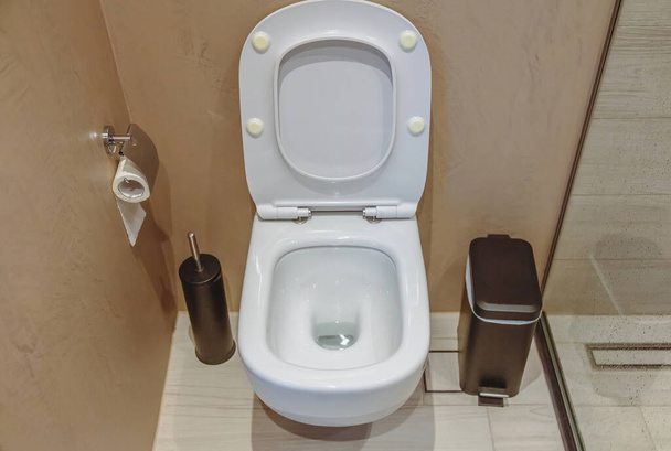 WC-sisustus, keraaminen valkoinen wc-kulho avoimella kannella ja mustalla harjalla ja roskakorilla. Moderni kylpyhuone tarvikkeet ruskeat seinät ja laatoitettu lattia tausta. - Valokuva, kuva