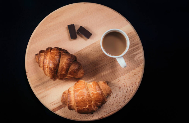 From above view of two fresh croissants, pieces of chocolate and  mug with coffee placed on napkin on black background of table - Foto, Imagem