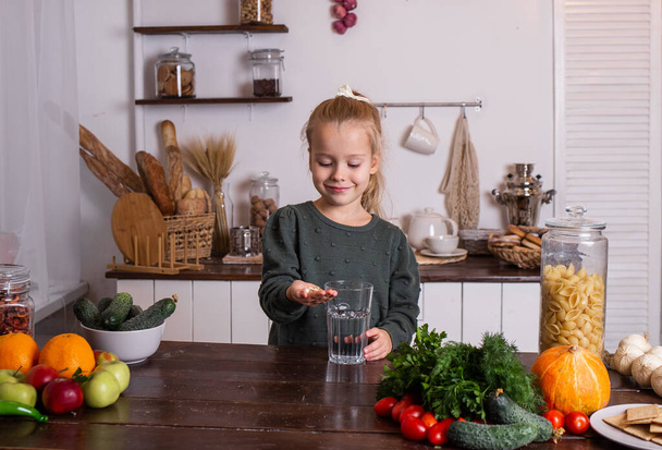 Egy szőke kislány ül a konyhaasztalnál és vitaminokat iszik. - Fotó, kép
