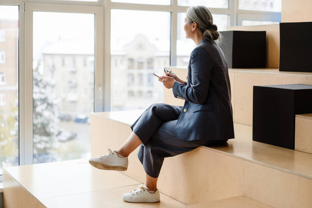 Elegant white-haired businesswoman using cellphone while sitting indoors - Foto, Bild