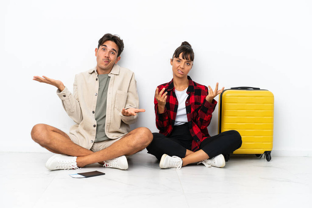 Jovens viajantes mestiços casal sentado no chão isolado no fundo branco infeliz e frustrado com algo porque não entender algo - Foto, Imagem