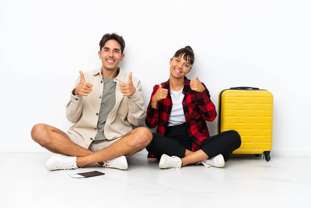 Jeune couple mixte de voyageurs assis sur le sol isolé sur fond blanc donnant un geste pouce levé avec les deux mains et souriant - Photo, image